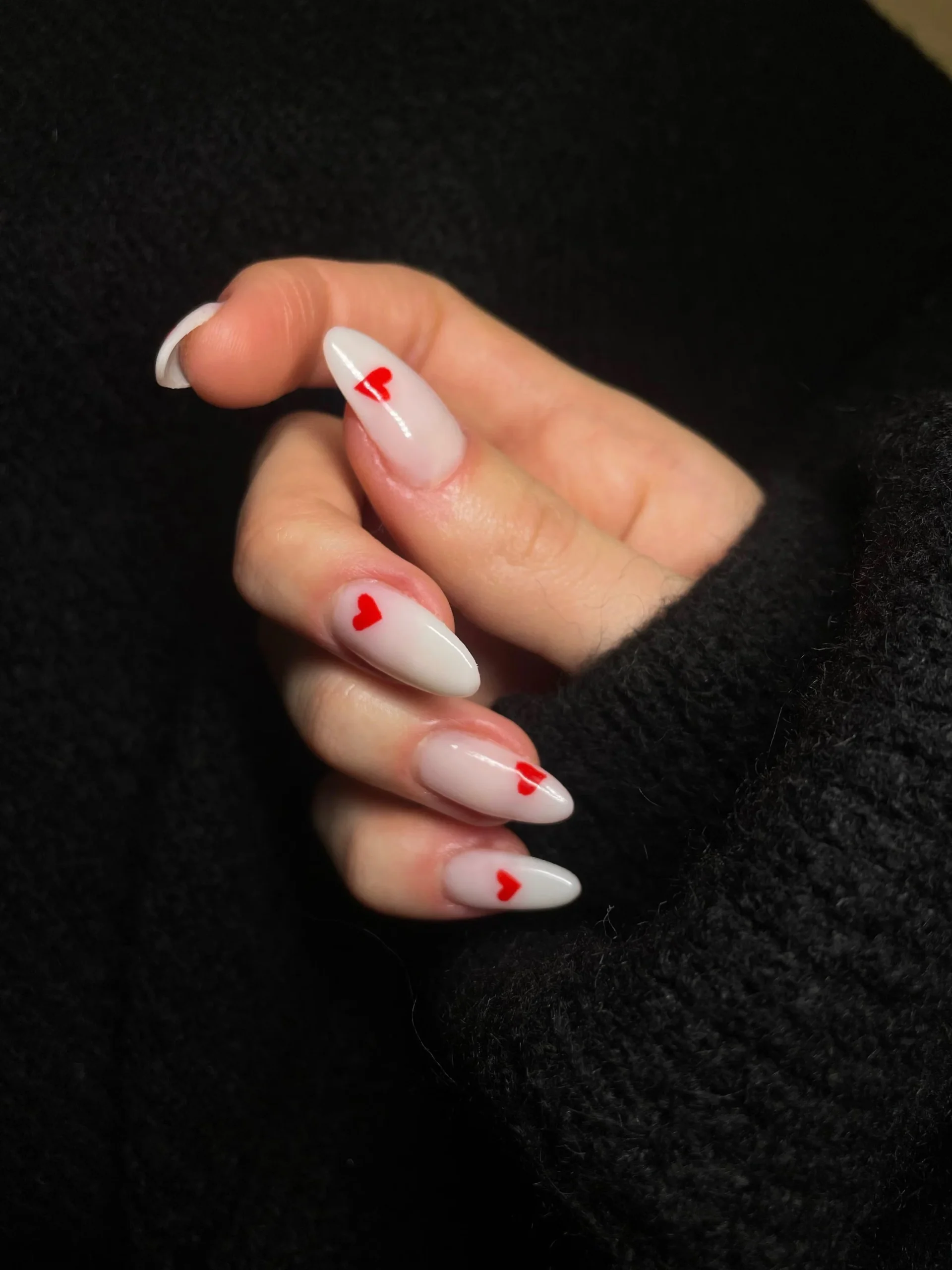 "Hand with white almond-shaped acrylic nails featuring small red heart nail art, against a black sweater background"